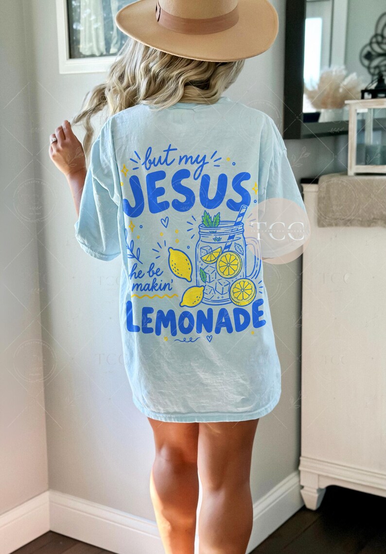 May include: A light blue short-sleeve t-shirt featuring a graphic on the back with the text "but my JESUS he be makin' LEMONADE". The design includes a blue mason jar filled with yellow lemon slices, ice cubes, and green mint leaves.