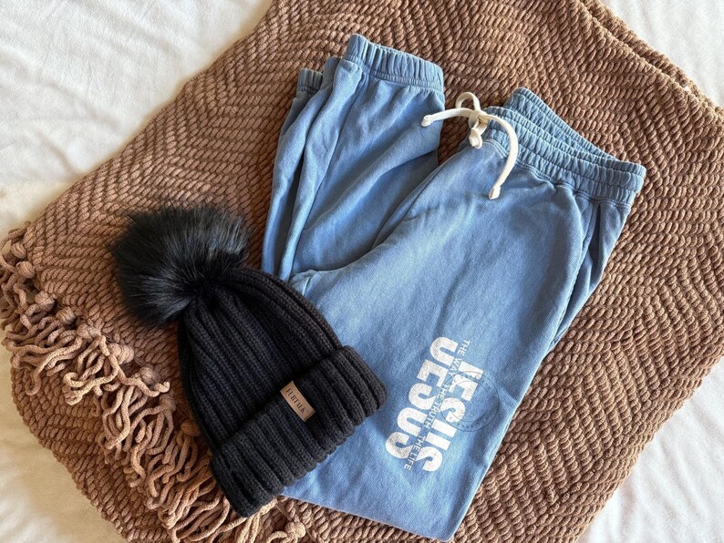 May include: A black knit beanie with a faux fur pom-pom and a small leather tag, paired with light blue sweatpants. The sweatpants have a drawstring and the text "JESUS THE WAY, THE TRUTH, THE LIFE" printed on the leg. The items are on a brown blanket.