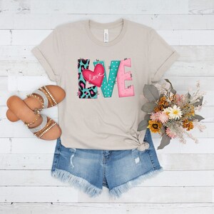 May include: A beige t-shirt with a graphic of the word "LOVE" in colorful patchwork letters. The letters are decorated with polka dots, animal print, and a pink heart with the word "love" inside.