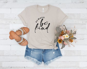 May include: A beige t-shirt with the text "ALWAYS be kind" in black lettering with a small heart symbol. The shirt is paired with denim shorts and sandals.