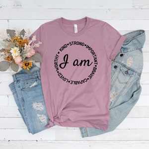 May include: A mauve t-shirt with a black circle graphic that says "I am" surrounded by the words "kind, strong, important, brave, capable, loved, worthy".
