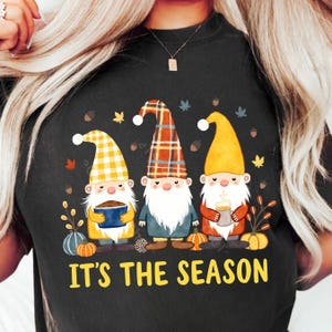 Puede incluir: Tres gnomos con sombreros de cuadros, amarillo y blanco a cuadros, sosteniendo un tazón, una taza y una taza. Los gnomos están rodeados de hojas de otoño y calabazas. El texto "Es la temporada" está escrito en letras amarillas debajo de los gnomos.