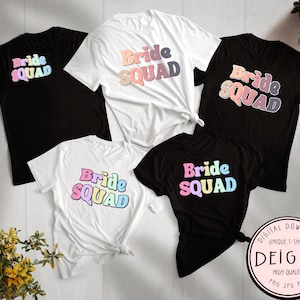 Puede incluir: Cinco camisetas negras y blancas con el texto "Bride Squad" en colores arcoíris. El texto está en un estilo de fuente retro. Las camisetas están dispuestas sobre una superficie blanca.