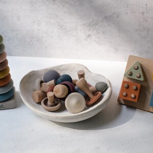 May include: A set of children's toys including a vertical stacking ring tower with multicolored rings, a white ceramic bowl filled with various small wooden mushroom-shaped pieces, and a wooden shape sorting board with geometric blocks in muted tones.