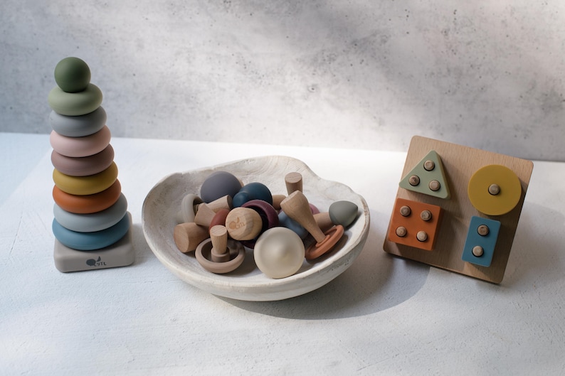 May include: A set of children's toys including a vertical stacking ring tower with multicolored rings, a white ceramic bowl filled with various small wooden mushroom-shaped pieces, and a wooden shape sorting board with geometric blocks in muted tones.