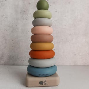 May include: A vertical stacking toy featuring a series of smooth, matte-finish rings in shades of olive green, sage, gray, blush, tan, mustard, burnt orange, and blue. The bottom blue ring reads I AM CONFIDENT, and the base features a logo with the text LYTL.