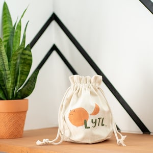 May include: A white drawstring bag with a cartoon orange cat design and the text "LYTL" printed on it. The bag is sitting on a wooden shelf with a green plant in a terracotta pot in the background.