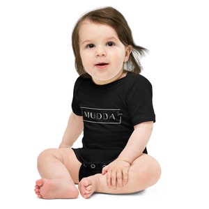 May include: A black baby onesie with white text that reads "MUDDA" in a box. A baby is wearing the onesie and sitting on a white background.