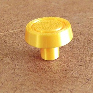 May include: A bright yellow cabinet knob with a rounded top and a cylindrical stem. The top has a textured, slightly raised center. The knob is made of a solid material and is set against a brown background.