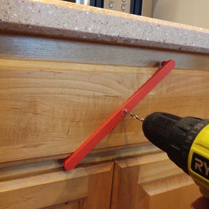 Peut inclure: Une poign&eacute;e de porte d'armoire rouge est perc&eacute;e dans une porte d'armoire en bois avec une perceuse Ryobi jaune et noire. L'armoire est brun clair et le comptoir est mouchet&eacute;.