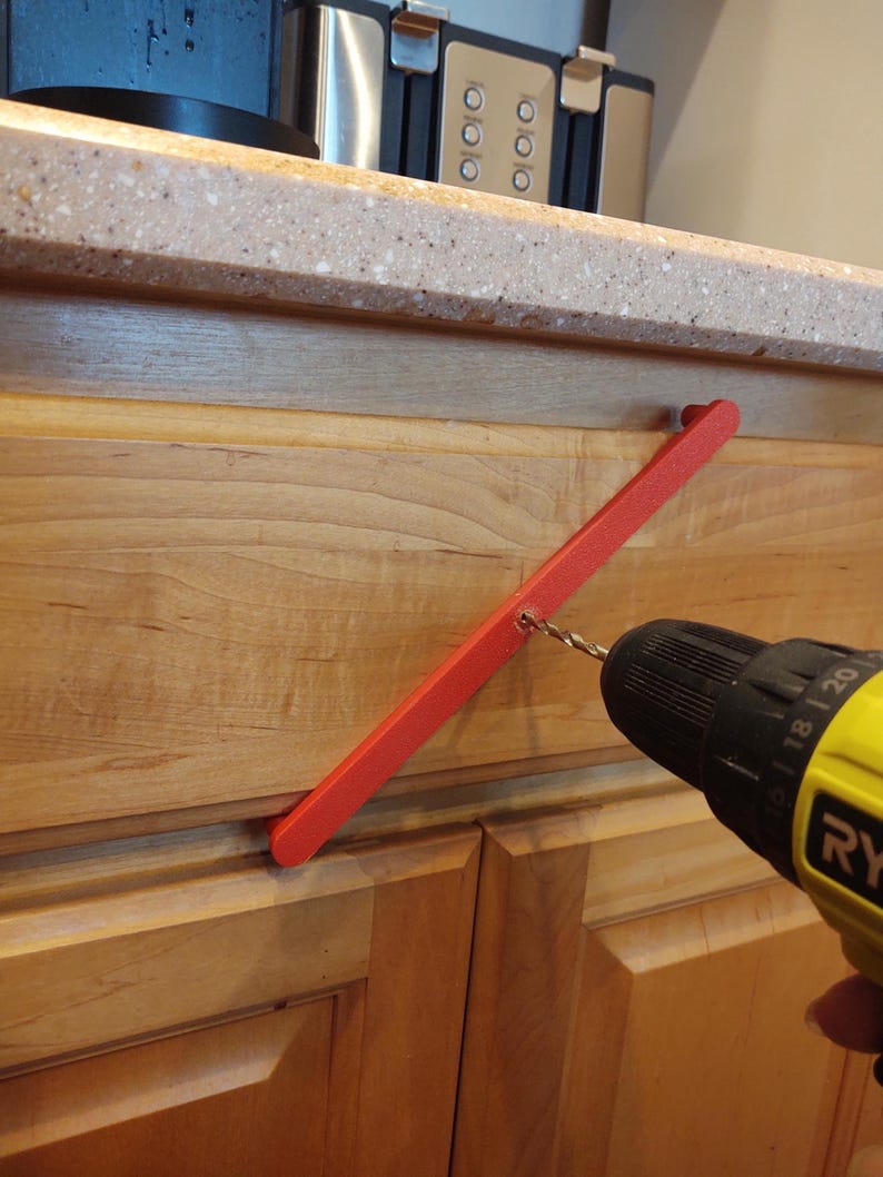 Peut inclure: Une poign&eacute;e de porte d'armoire rouge est perc&eacute;e dans une porte d'armoire en bois avec une perceuse Ryobi jaune et noire. L'armoire est brun clair et le comptoir est mouchet&eacute;.