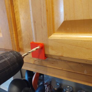 Peut inclure: Un guide de per&ccedil;age rouge est fix&eacute; &agrave; une porte d'armoire en bois. Un foret est ins&eacute;r&eacute; dans le guide, pr&ecirc;t &agrave; faire un trou. L'armoire est en bois de couleur claire.