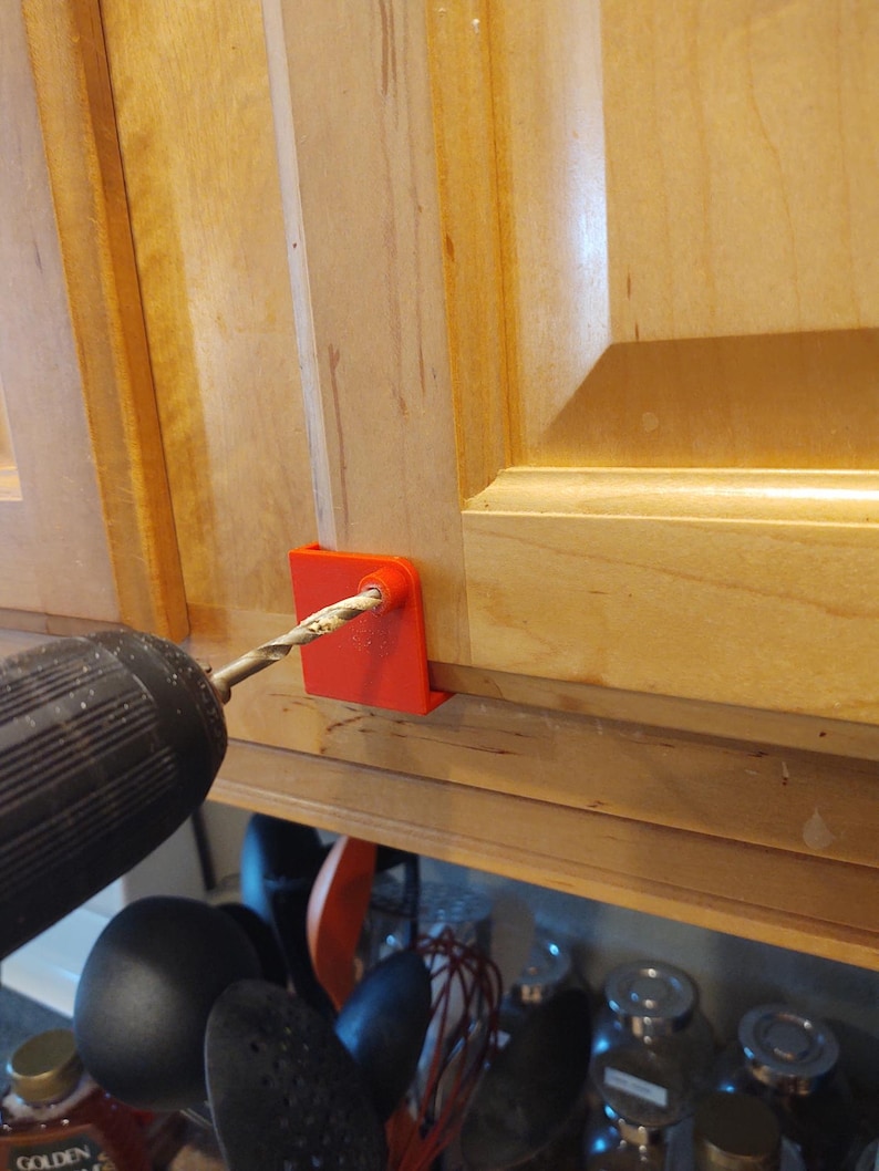 Peut inclure: Un guide de per&ccedil;age rouge est fix&eacute; &agrave; une porte d'armoire en bois. Un foret est ins&eacute;r&eacute; dans le guide, pr&ecirc;t &agrave; faire un trou. L'armoire est en bois de couleur claire.