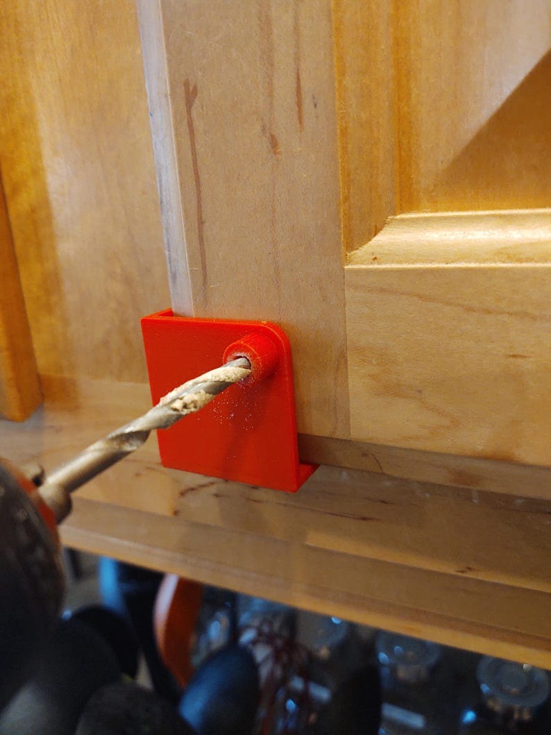 Peut inclure: Un guide de per&ccedil;age rouge vif est serr&eacute; sur une porte d'armoire en bois. Un foret est ins&eacute;r&eacute; dans le guide, cr&eacute;ant un trou dans le bois. La porte de l'armoire est brun clair et la perceuse est noire.