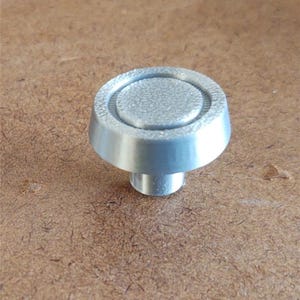 Pode incluir: Um puxador de armário prateado com um design circular. O puxador tem um aspeto texturizado, ligeiramente fosco na superfície superior e uma base cónica lisa. O puxador tem aproximadamente 2,5 cm de diâmetro.