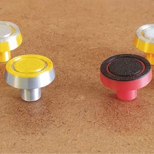 May include: A collection of colorful knobs with a circular design. The knobs feature a variety of colors, including red, yellow, blue, and silver. The knobs are arranged on a brown surface.