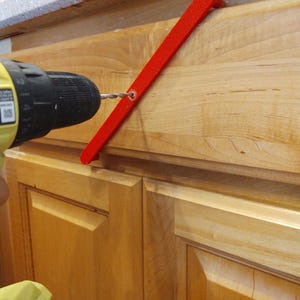 Peut inclure: Un guide d'installation de quincaillerie d'armoire rouge vif est utilis&eacute; avec une perceuse jaune et noire pour installer une poign&eacute;e d'armoire. Le guide est positionn&eacute; contre une porte d'armoire en bois clair.