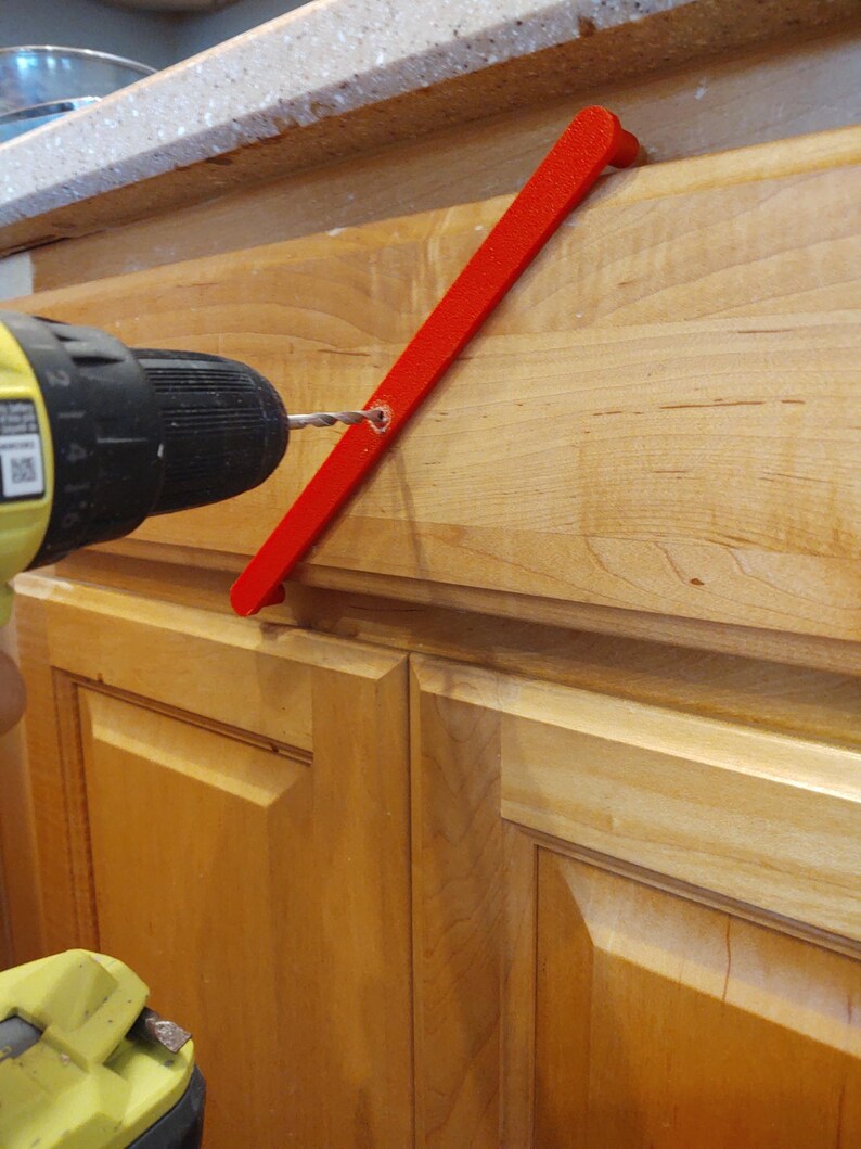 Peut inclure: Un guide d'installation de quincaillerie d'armoire rouge vif est utilis&eacute; avec une perceuse jaune et noire pour installer une poign&eacute;e d'armoire. Le guide est positionn&eacute; contre une porte d'armoire en bois clair.