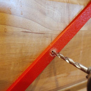 Peut inclure: Gros plan d'une perceuse per&ccedil;ant un trou dans une barre m&eacute;tallique rouge vif textur&eacute;e. La barre est inclin&eacute;e contre une surface en bois clair, sugg&eacute;rant un projet de bricolage ou de construction.