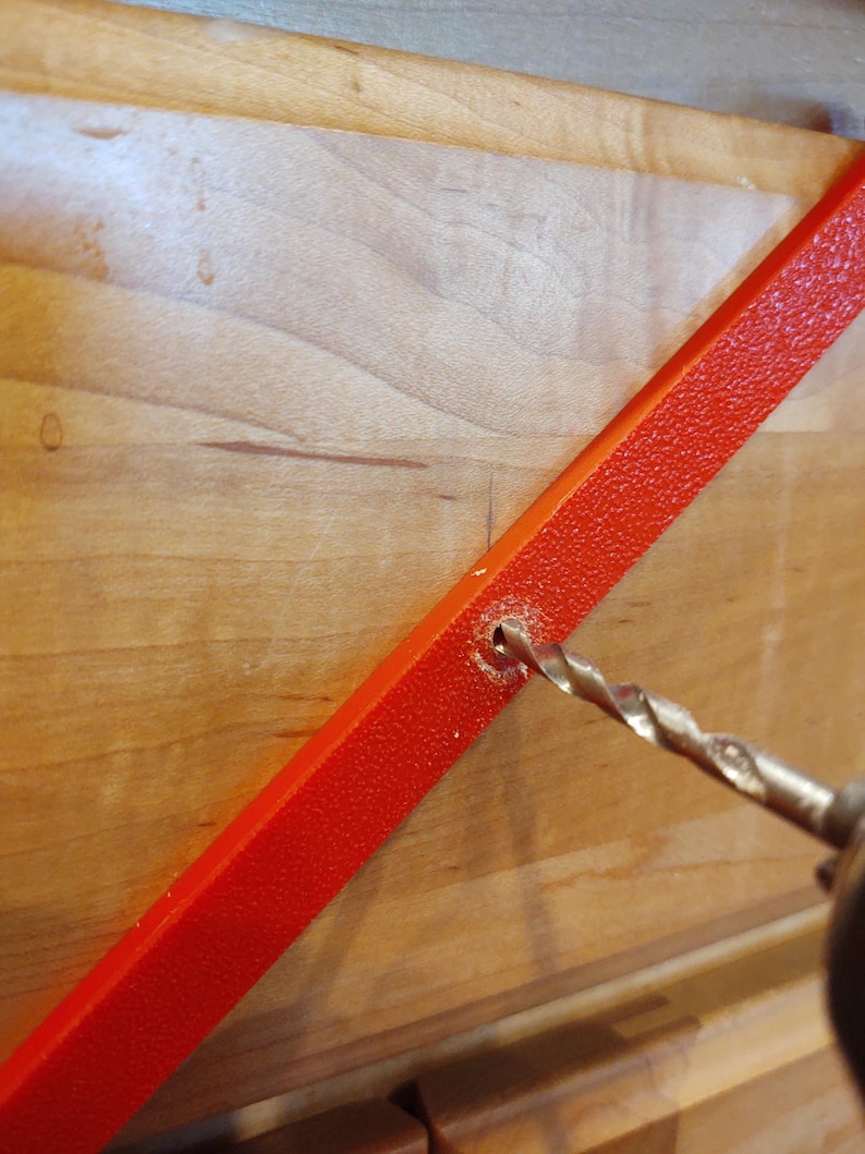 Peut inclure: Gros plan d'une perceuse per&ccedil;ant un trou dans une barre m&eacute;tallique rouge vif textur&eacute;e. La barre est inclin&eacute;e contre une surface en bois clair, sugg&eacute;rant un projet de bricolage ou de construction.