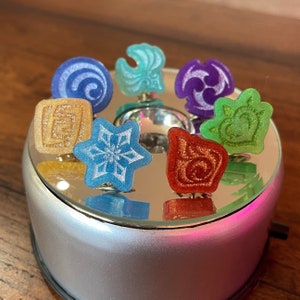 May include: A set of eight colorful enamel pins featuring different designs. The pins are arranged in a circle on a silver display stand. The designs include a snowflake, a heart, a star, a swirl, and a few abstract shapes.
