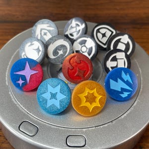 May include: A collection of 12 round, colorful, and textured tokens with various symbols, including stars, lightning bolts, and a fire symbol. The tokens are arranged on a silver surface.