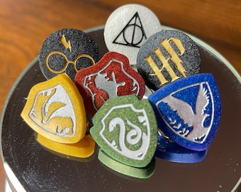 May include: Seven different Harry Potter house crests, each with a different color and design. The crests are made of metal and have a shiny finish. The crests are arranged in a circle on a black surface.