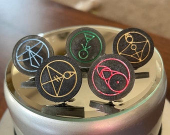 May include: Five black circular pins with different colored and gold designs. The pins are on a silver surface.