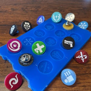 May include: A blue silicone mold with 16 circular cavities for making resin pins. The mold is filled with various colorful resin pins with different designs, including a snowflake, a swirl, a plus sign, a gun, and a sword.