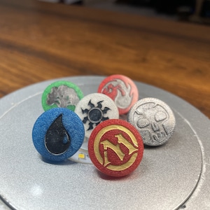 May include: Six round, colorful, plastic tokens with different symbols. The tokens are on a silver surface. The symbols include a tree, a sun, a skull, a drop of water, a flame, and a letter "M".
