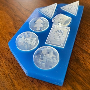 May include: A blue silicone mold with eight clear plastic molds for making resin crafts. The molds feature various shapes, including a triangle, a circle, and a rectangle. The molds are filled with a clear liquid, which is likely resin.