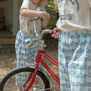 May include: Two children wearing matching outfits stand near a red bicycle. The outfits include white t-shirts with a logo and blue pants patterned with white swans. One child wears a red and white checkered hat.