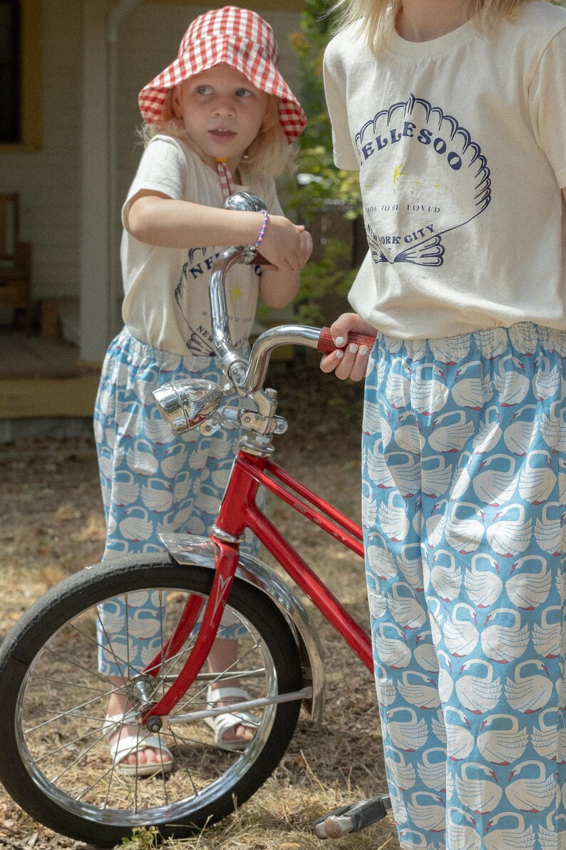 May include: Two children wearing matching outfits stand near a red bicycle. The outfits include white t-shirts with a logo and blue pants patterned with white swans. One child wears a red and white checkered hat.