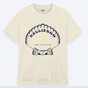 May include: Cream-colored t-shirt featuring a navy blue seashell design with the text "NELLIESOO" and "MADE TO BE LOVED" and "NEW YORK CITY EST 2023". The shirt has a classic crew neck and short sleeves.