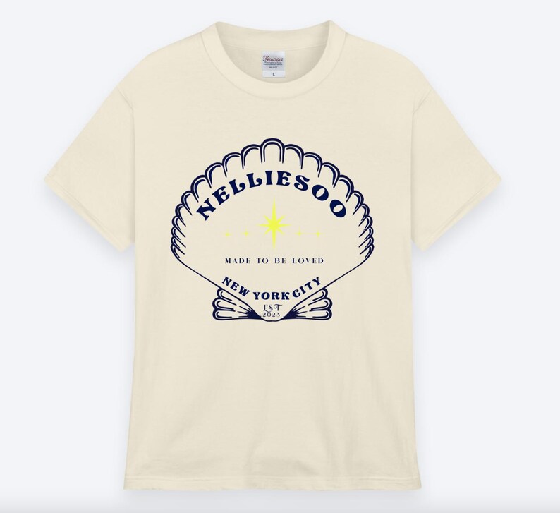 May include: Cream-colored t-shirt featuring a navy blue seashell design with the text "NELLIESOO" and "MADE TO BE LOVED" and "NEW YORK CITY EST 2023". The shirt has a classic crew neck and short sleeves.