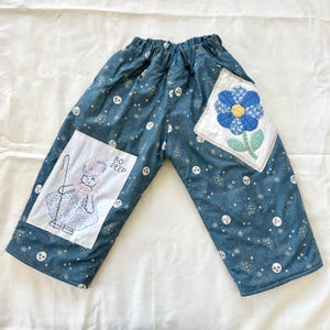 Op de afbeelding: Blauwgepatenteerde broek met elastische tailleband. De broek heeft geborduurde patches, een met een bloem en de andere met een cartoonillustratie van Bo Peep en de tekst "BO PEEP". De broek is versierd met witte sterren en lachende maan gezichten.