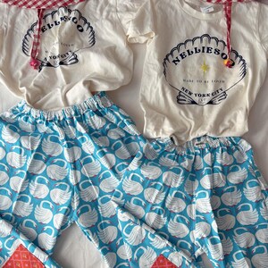 May include: Matching children's clothing set featuring a white t-shirt with a seashell graphic and the text "NELLIESO" and "MADE TO BE LOVED NEW YORK CITY". The set includes blue swan print pants and a red gingham hat.