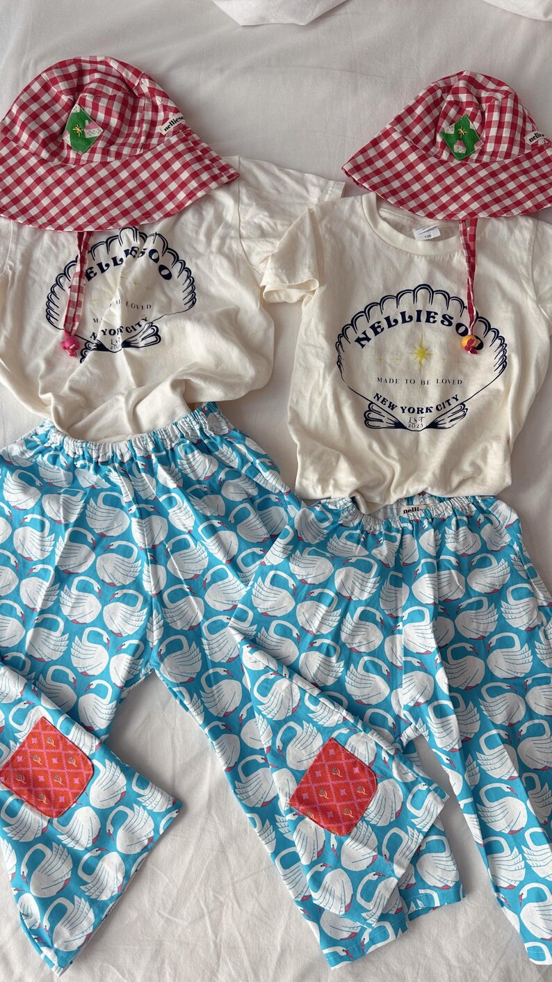 May include: Matching children's clothing set featuring a white t-shirt with a seashell graphic and the text "NELLIESO" and "MADE TO BE LOVED NEW YORK CITY". The set includes blue swan print pants and a red gingham hat.