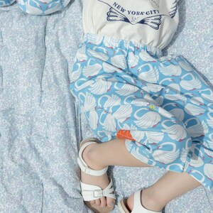 May include: A child's outfit featuring a white t-shirt with a seashell design and the text "NELLIESOO NEW YORK CITY". The outfit includes blue pants with a white swan pattern and white sandals.