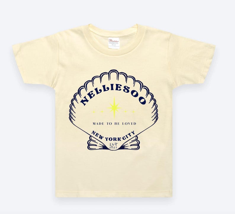 May include: Cream-colored t-shirt featuring a navy blue seashell design with the text "NELLIESOO" and "MADE TO BE LOVED" and "NEW YORK CITY LST 2023". The design includes a yellow star.