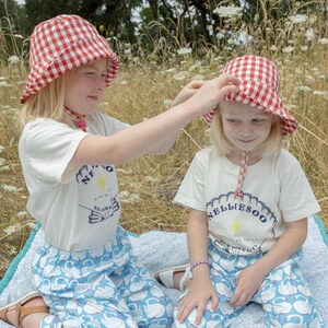 May include: Two young children wearing matching outfits, including red and white gingham bucket hats, white t-shirts with a graphic, and blue pants with a swan pattern. They are sitting on a blue and white patterned blanket.