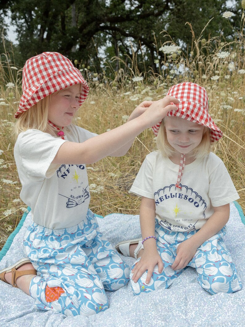 May include: Two young children wearing matching outfits, including red and white gingham bucket hats, white t-shirts with a graphic, and blue pants with a swan pattern. They are sitting on a blue and white patterned blanket.