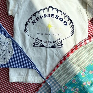 May include: Cream-colored t-shirt with a seashell design and the text "Nelliesoo, Made to be Loved, New York City, Est 2023." A blue and white gingham banner and a patchwork fabric with floral and striped patterns are also visible.