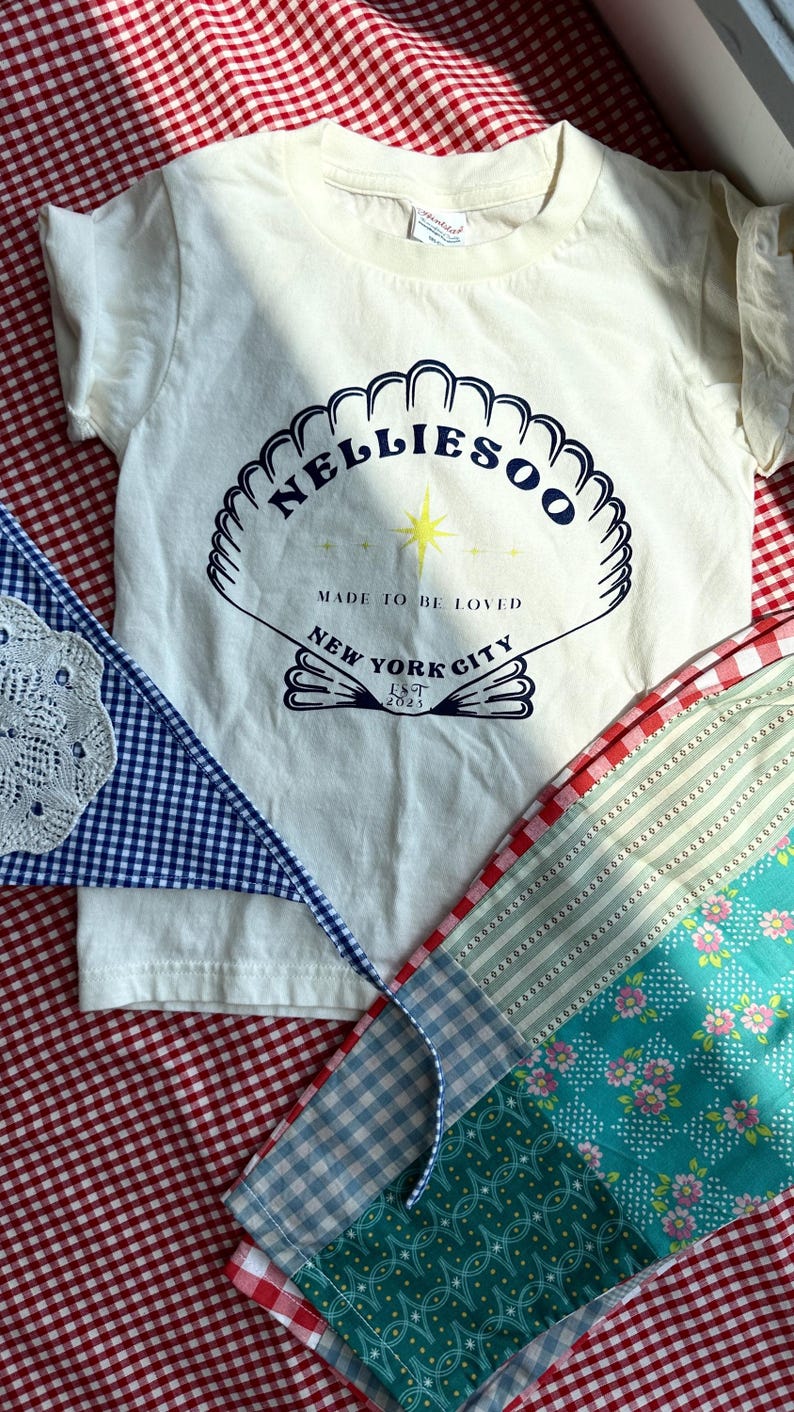May include: Cream-colored t-shirt with a seashell design and the text "Nelliesoo, Made to be Loved, New York City, Est 2023." A blue and white gingham banner and a patchwork fabric with floral and striped patterns are also visible.