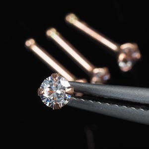 Könnte beinhalten: Nahaufnahme eines funkelnden, rund geschliffenen Diamantohrsteckers, der von einer Pinzette gehalten wird, mit drei ähnlichen Ohrringen im Hintergrund. Die Ohrringe haben eine Roségoldfassung und einen klaren, brillanten Edelstein. Der Hintergrund ist schwarz.