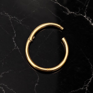 May include: A circular gold hoop ring featuring a hinged segment closure mechanism. The metal surface has a polished finish and the engraving 9K is clearly visible on the lower portion of the hoop, indicating the gold purity of the jewelry piece.