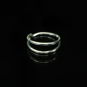 Puede incluir: Un aro de nariz plateado de doble aro. El anillo está hecho de un metal brillante y tiene un diseño simple y minimalista. El anillo se muestra sobre un fondo negro.