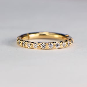 May include: A gold band ring with a continuous row of small, clear gemstones set around the circumference. The ring is made of polished gold and the gemstones sparkle against the gold setting. The ring is displayed on a white surface.