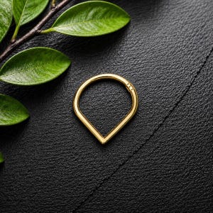 May include: A gold-toned teardrop-shaped hoop ring with a smooth, polished finish. The jewelry piece features a continuous circular top that tapers into a pointed bottom, creating a distinct geometric silhouette. The metal surface is reflective.