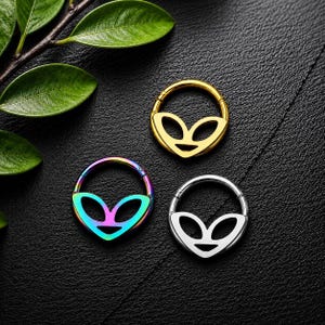 May include: Three circular hinged hoop rings featuring a cutout alien head design. One ring is gold-toned, one is silver-toned, and one features an iridescent rainbow finish. Each ring has a smooth, polished surface and a secure clicker closure mechanism.